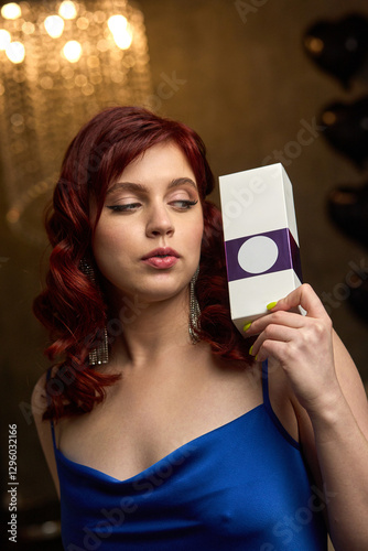 Obraz na płótnie Elegant redhead woman in blue dress holding a white box with purple label and thoughtful expression, glamour concept w salonie