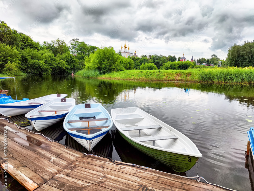 Obraz premium Boats lined up by the tranquil river under cloudy skies near a historic site