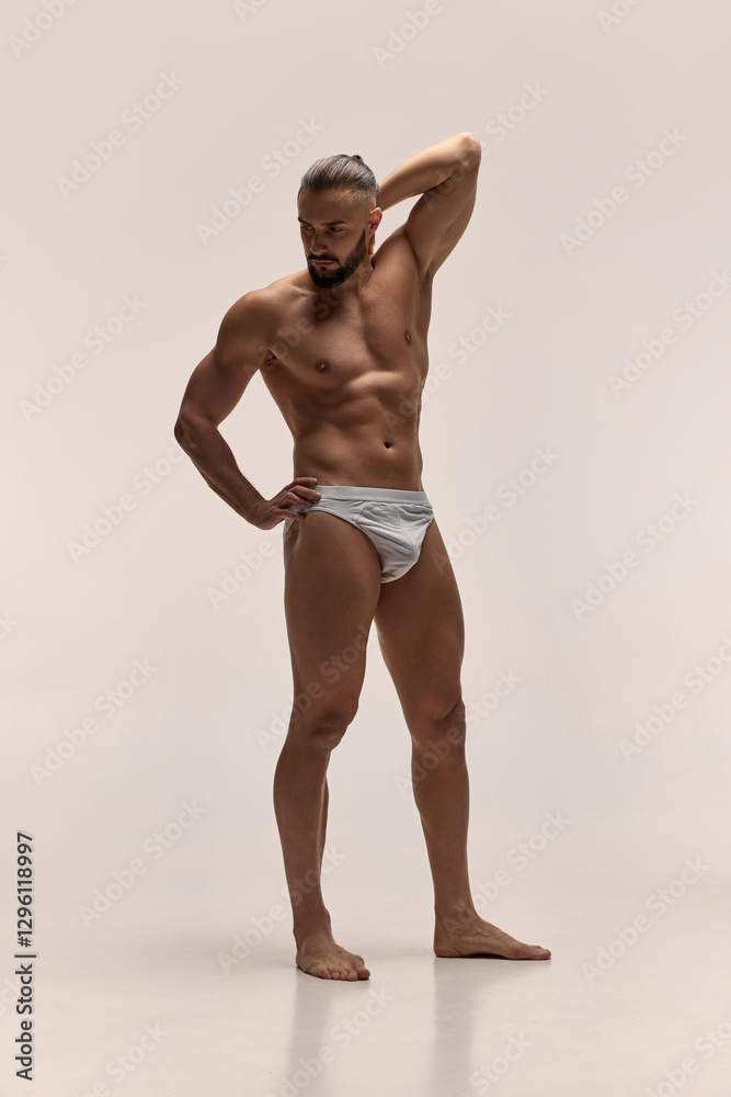 Fototapeta premium Full-length portrait of handsome man with muscular strong body posing shirtless in underwear against light studio background. Concept of male beauty, healthy and supportive lifestyle, wellness