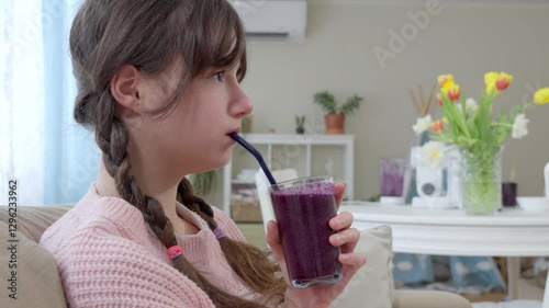 A teenage girl in a pink knitted sweater is drinking a freshly blended berry smoothie from a glass. Healthy nutrition, homemade drinks, and a balanced diet for kids and teenagers