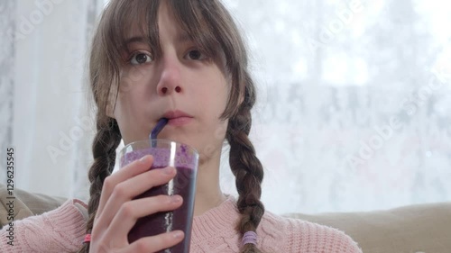 A teenage girl in a pink knitted sweater is drinking a freshly blended berry smoothie from a glass. Healthy nutrition, homemade drinks, and a balanced diet for kids and teenagers