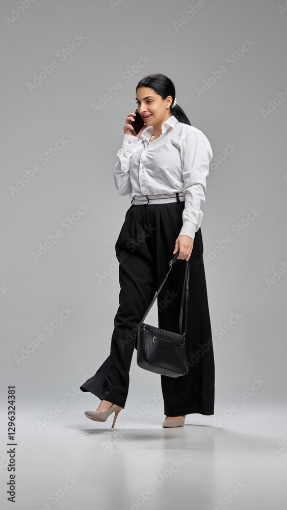 Full-length portrait of positive young business woman in formal wear walking and talking on smartphone against grey studio background. Concept of communication, human emotions, lifestyle Full-length portrait of positive young business woman in formal wear walking and talking on smartphone against grey studio background. Concept of communication, human emotions, lifestyle
