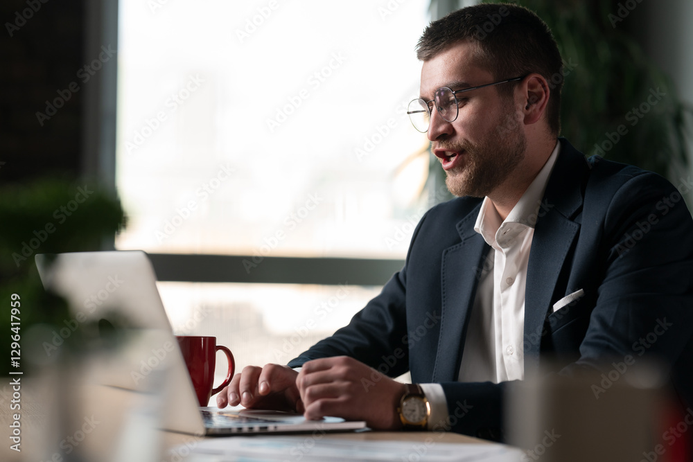 Obraz premium Young businessman in a suit and glasses working on a laptop in a modern office. Concept of professional communication, remote work, online meeting, corporate lifestyle, entrepreneurship