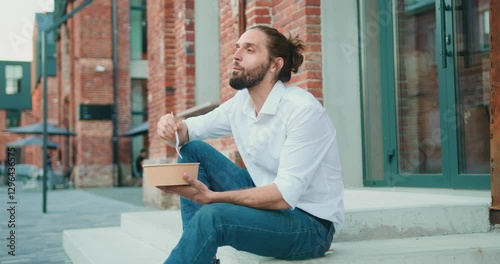 Business man in white shirt eating healthy salad on a break sitting on stairs over office building background. Male dieting nutrition concept