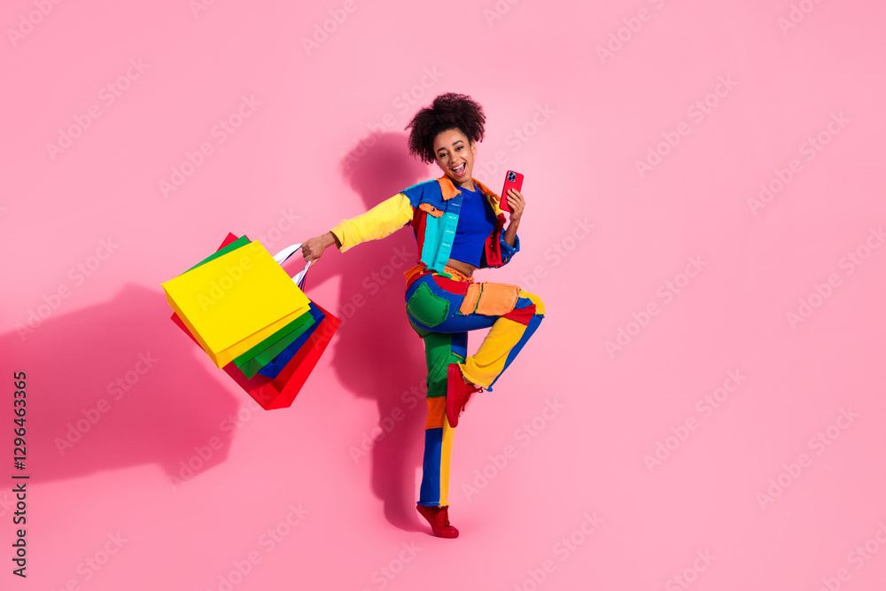 Fototapeta premium Stylish young woman in colorful outfit holding shopping bags on pink background with joyful expression