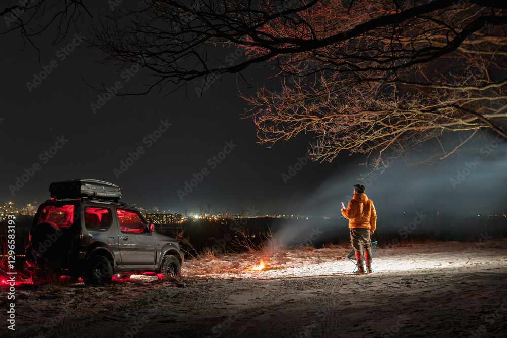A man stands with a lantern on his head. lights of the night city on background. A bonfire burns with smoke. An SUV vehicle is parked on top. The concept of recreation and trips to nature.