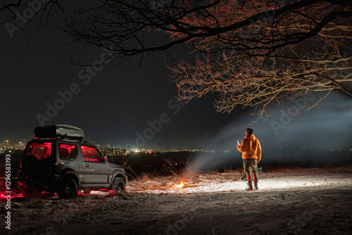 Obraz na płótnie A man stands with a lantern on his head. lights of the night city on background. A bonfire burns with smoke. An SUV vehicle is parked on top. The concept of recreation and trips to nature. w salonie