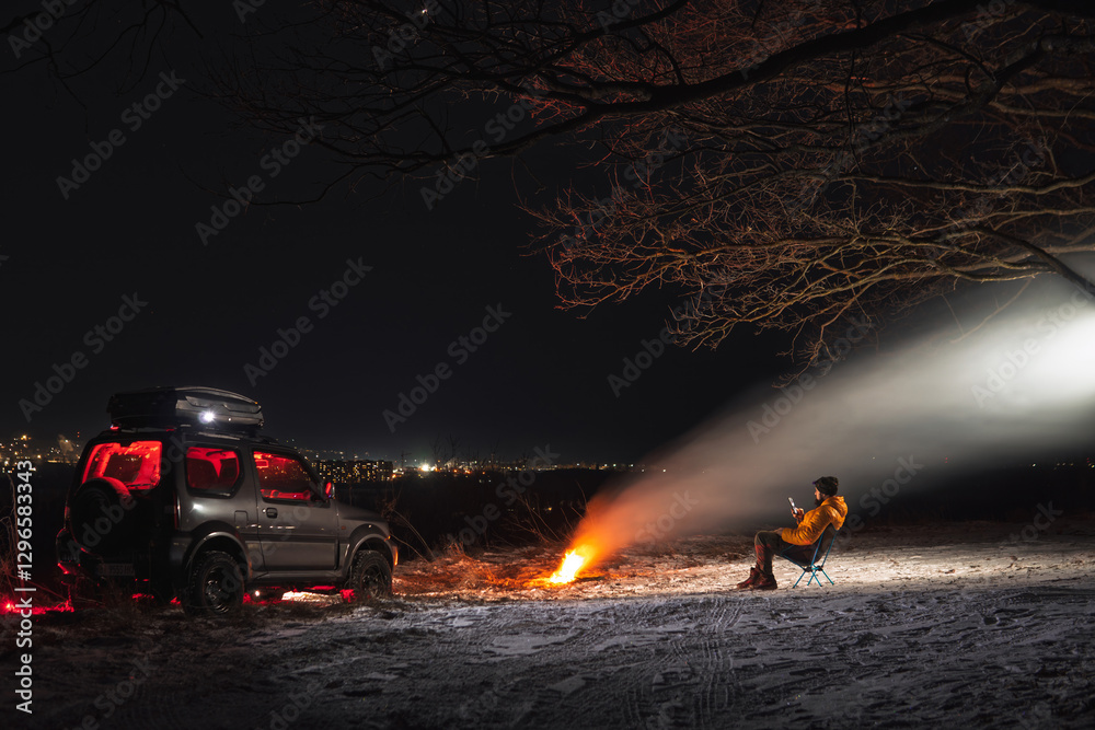 A man sits on a chair with a view of the lights of the night city. A bonfire burns with smoke. An off-road vehicle is parked on top. The concept of recreation and trips to nature.