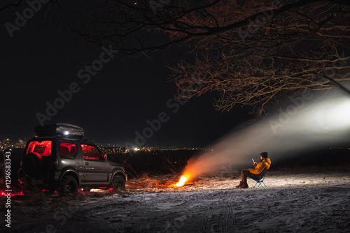 Obraz na płótnie A man sits on a chair with a view of the lights of the night city. A bonfire burns with smoke. An off-road vehicle is parked on top. The concept of recreation and trips to nature. w salonie