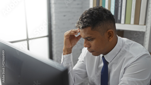Young, hispanic man in an office wearing a white shirt and blue tie, focusing intently on computer work near a window with shelves in the background.