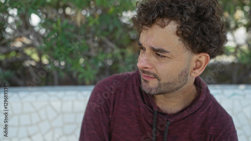 Young hispanic man with curly hair in a maroon hoodie sitting on a mosaic bench in an urban park, looking contemplatively towards the lush greenery around.