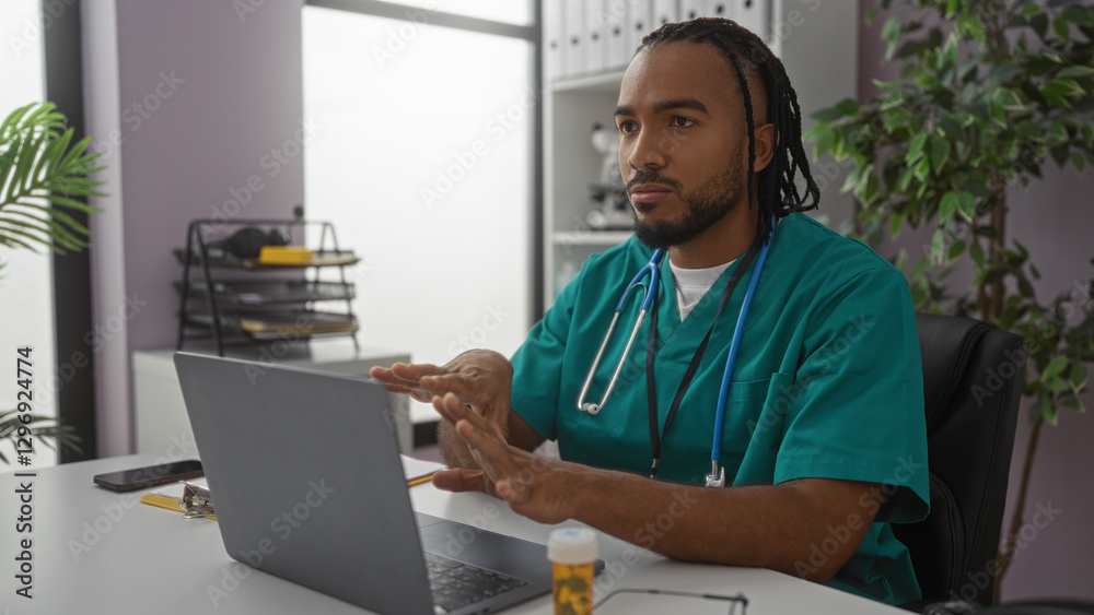 Fototapeta premium Young man with braids working on a laptop in a clinic room wearing scrubs with stethoscope around neck and papers on desk in indoor medical workplace setting.