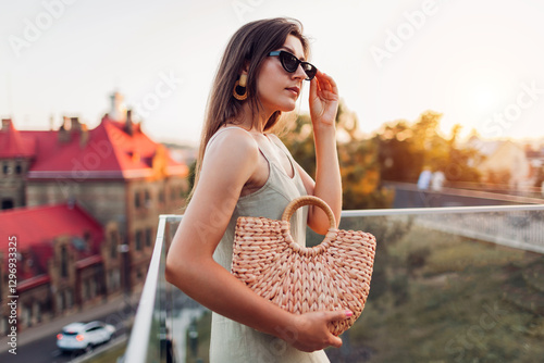 Portrait of young fashionable woman wearing summer outfit on city street in evening. Stylish straw wicket handbag