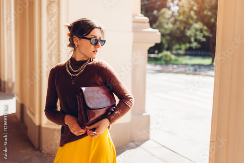 Portrait of young woman wears brown cashmere sweater and yellow skirt outfit on city street with stylish leather handbag