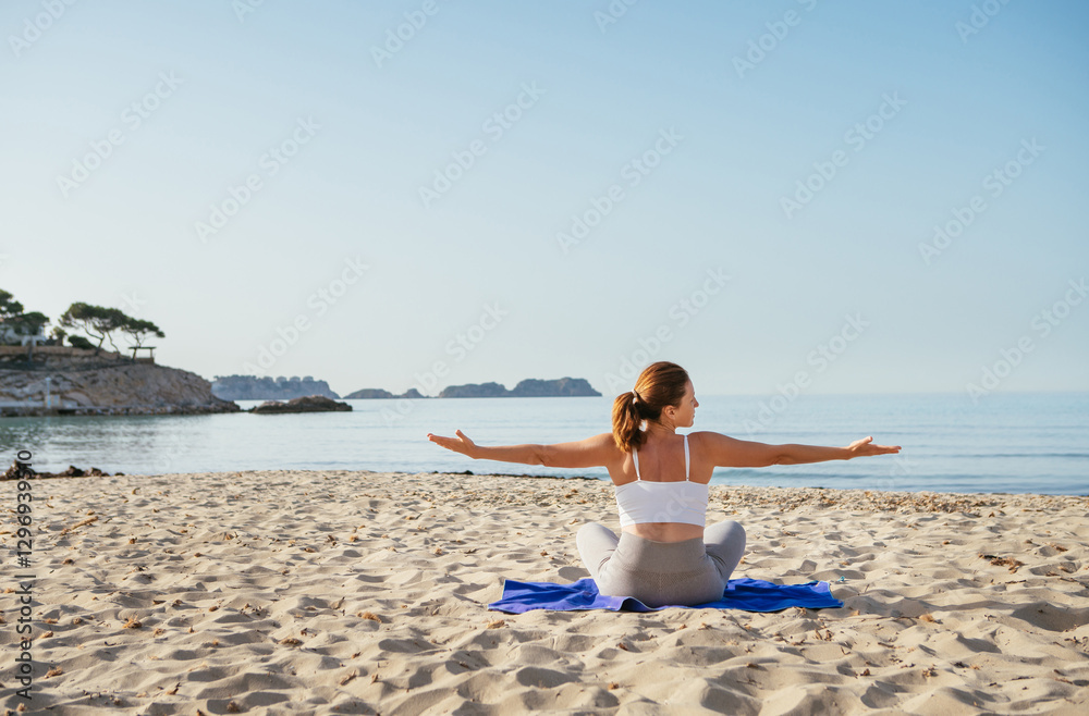 Fototapeta premium Calm woman sitting and doing deep breathing and stratching exercises and meditating in early morning hours on sandy beach with calm sea waves. Mental health, people relaxing, traveling concept.