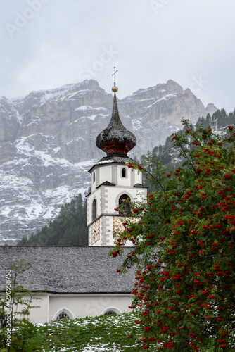 Chiesa di San Vigilio in Colfosco, with its distinctive onion dome, stands amid a wintry scene of fresh snow and red rowan berries, backed by rugged Dolomite mountains photo
