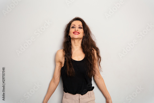 Positive relaxing smiling make-up woman with red lipstick and long natural curly hair style looking front in black summer top on blue studio empty copy space background. Expression portrait