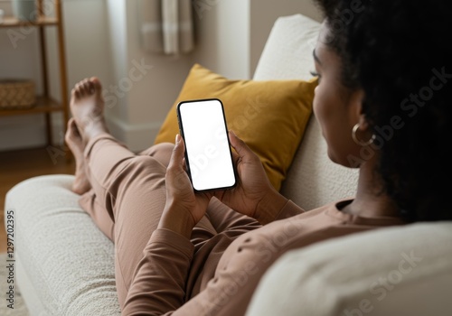 Unrecognizable woman holding a smartphone with a white screen at home, over the shoulder view of lady using a blank mobile phone while relaxing on a couch.

