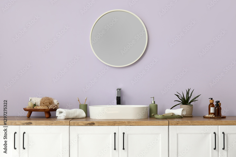 Obraz premium Mirror, chest of drawers with sink and bath supplies near lilac wall in interior of bathroom