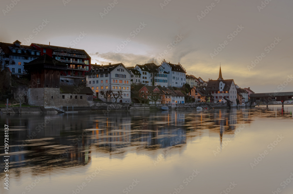 Naklejka premium Reflections of historic buildings along the calm river at sunset in a tranquil town