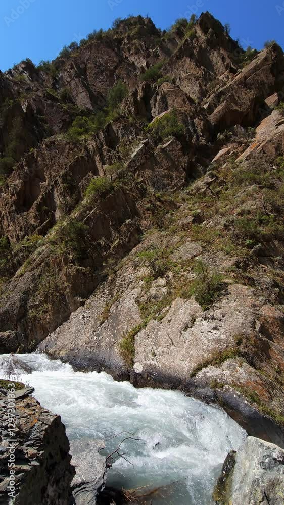 Mountain River Flowing Through at the foot of large rocky mountain cliff at sunny summer day Mountain River Flowing Through at the foot of large rocky mountain cliff at sunny summer day