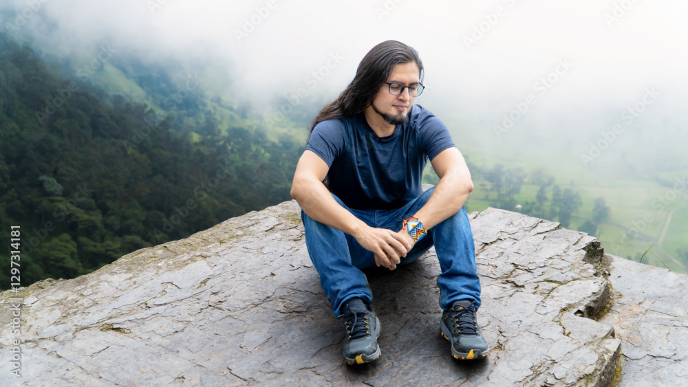 Naklejka premium Smiling young man sits on a rocky cliff with a misty green valley in the background. Peaceful outdoor adventure in nature