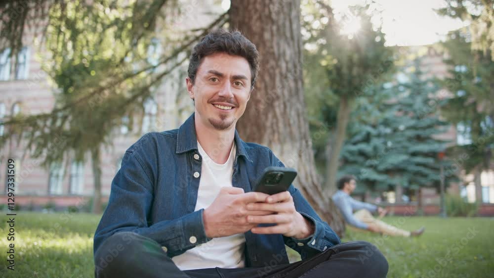 Attractive Caucasian guy sitting in lotus position on grass at park of campus. Using smartphone for remote communication. Positively looking at camera. In background other students passing by. Attractive Caucasian guy sitting in lotus position on grass at park of campus. Using smartphone for remote communication. Positively looking at camera. In background other students passing by.