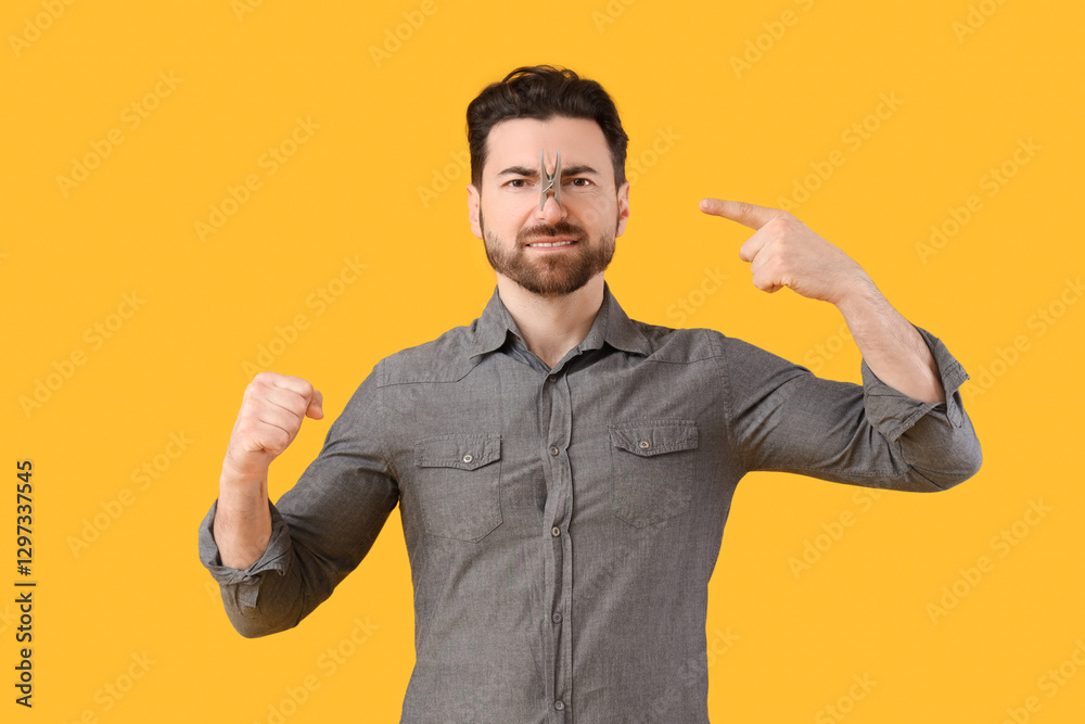 Ill young man pointing at clothespin on his nose against yellow background. Stuffy nose concept Ill young man pointing at clothespin on his nose against yellow background. Stuffy nose concept