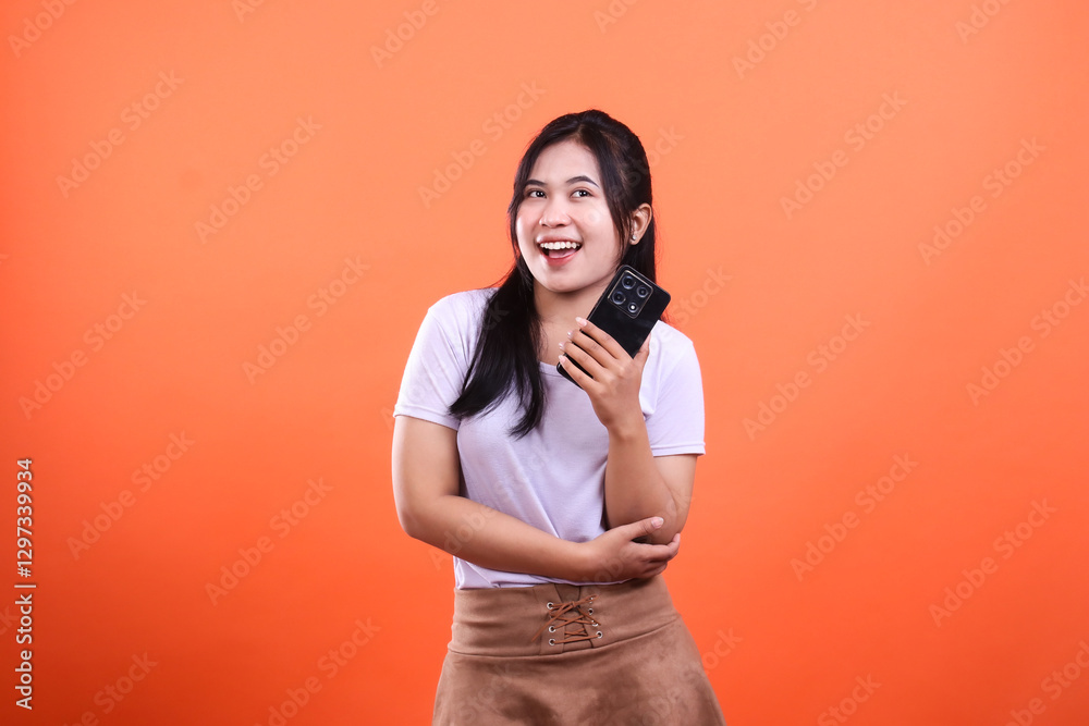 A woman with long black hair smiles holding a cellphone against an orange background A woman with long black hair smiles holding a cellphone against an orange background