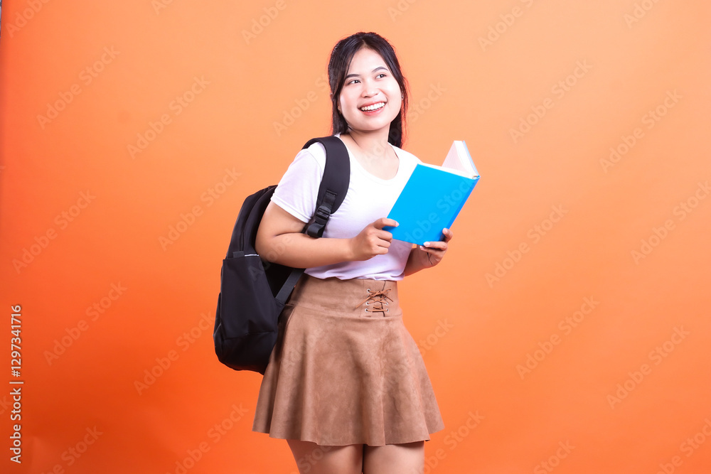 A smiling student holding a blue book and carrying a black backpack is looking at something in front of her A smiling student holding a blue book and carrying a black backpack is looking at something in front of her