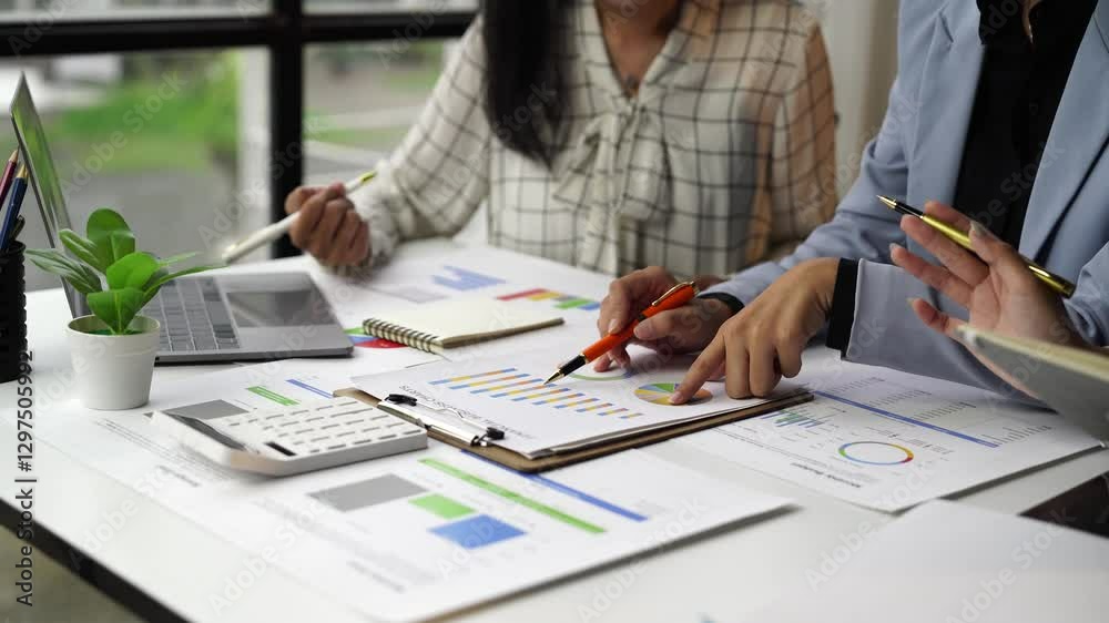 Two businesswomen are examining financial charts, pointing at graphs and pie charts with pens, having a discussion about data and using a calculator and laptop for analysis Two businesswomen are examining financial charts, pointing at graphs and pie charts with pens, having a discussion about data and using a calculator and laptop for analysis