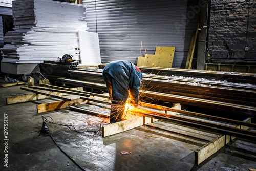 a worker in protective clothing, engaged in welding metal structures. Sparks fly from the welding site, illuminating the work area. There are various materials and details around, such as metal beams