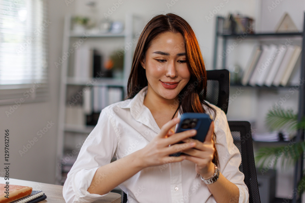 Smiling asian woman looking at smartphone screen, sitting on desk, enjoying leisure time with mobile device at home, happy young female chatting in social network or surfing internet, shopping online. Smiling asian woman looking at smartphone screen, sitting on desk, enjoying leisure time with mobile device at home, happy young female chatting in social network or surfing internet, shopping online.