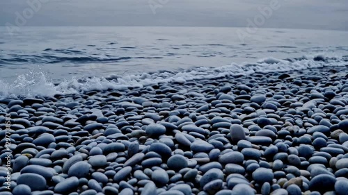 Static wide shot: scattered pebbles as waves roll in layers beyond, the cool tone color grade enhancing the tranquil mood.
