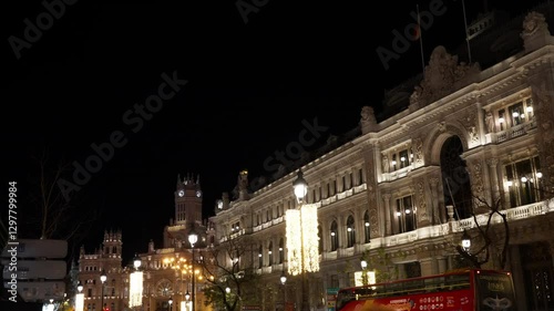 A historic ornate building lit by nighttime lights, with towers in the background and a passing bus in the foreground, under a black sky in central Madrid photo