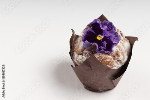Side view of a muffin dusted with powdered sugar, wrapped in brown parchment, and adorned with a deep purple edible flower.