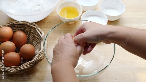 Hands Separating Egg Yolk from Egg White into Glass Bowl on Wooden Kitchen Counter with Basket of Eggs and Baking Ingredients in Background