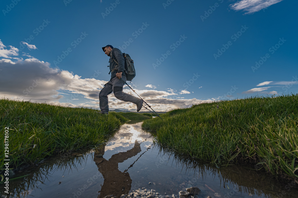 Obraz premium A tourist traveler with a backpack on his back and trekking poles jumps over a stream with green grass. A man is reflected in the water during a jump. Photo from the side of the water jump.