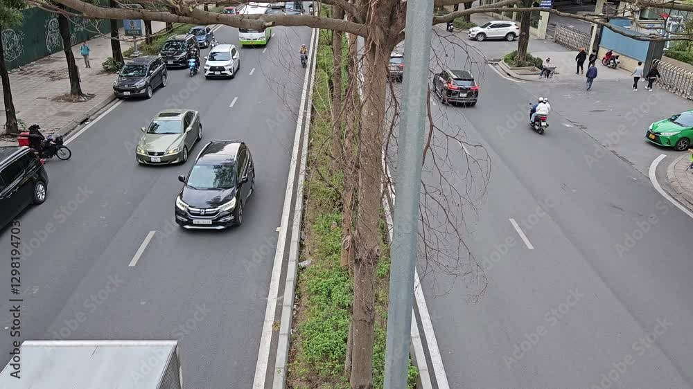 Aerial view of a bustling urban street scene, featuring moving vehicles, motorbikes, and pedestrians, with modern high-rise buildings and green trees lining the road in Ha Noi city, Vietnam.