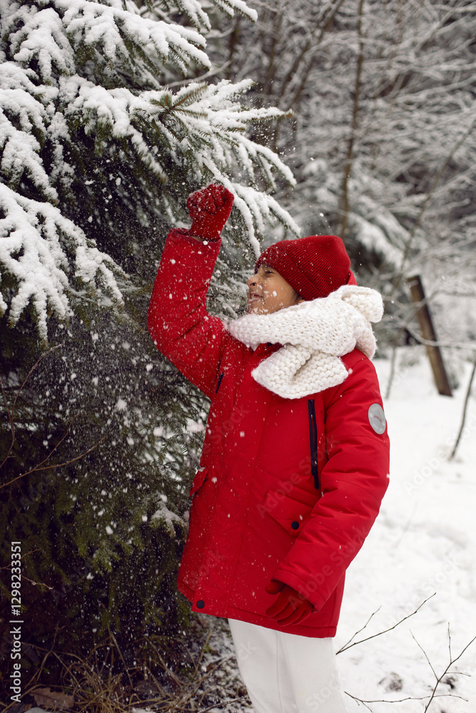 A cheerful seven-year-old boy in a red jacket walks outside in the winter in the forest and plays with snow during the day in clear weather
