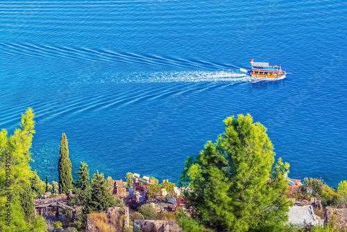A boat sails through the turquoise waters of the Mediterranean Sea near Alanya Castle, Turkey, leaving behind a trail of ripples. The lush green trees on the cliffside add to the scenic view