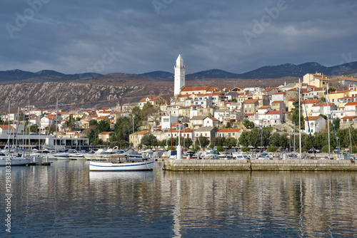 Novi Vinodolski, Croatia, presents a picturesque harbor with a white church spire overlooking the town. Boats rest on calm waters, reflecting the charming hillside architecture under a cloudy sky.