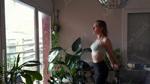 CHICA HACIENDO EJERCICIOS EN CASA. CÁMARA LENTA DE MUJER CON ROPA DEPORTIVA HACIENDO ESTIRAMIENTOS O YOGA RODEADA DE PLANTAS CON LUZ NATURAL. BARRIDO DE CÁMARA.