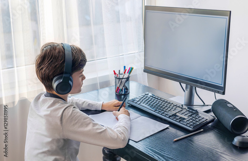 A child aged 7-9 sits at home behind a computer, writing assignments in a notebook. The image captures remote learning, digital study routines,  challenges associated with autism and ADHD. copyspace photo