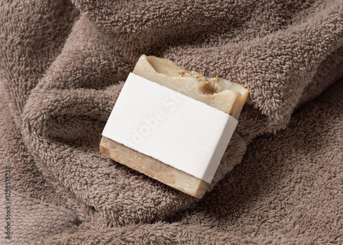Beige herbal soap bar with blank label on light brown bath towel top view, hygiene product mockup