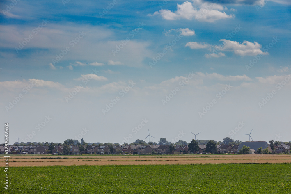 Fototapeta premium Expansive farmland under a cloudy sky with wind turbines in the distance near a small village