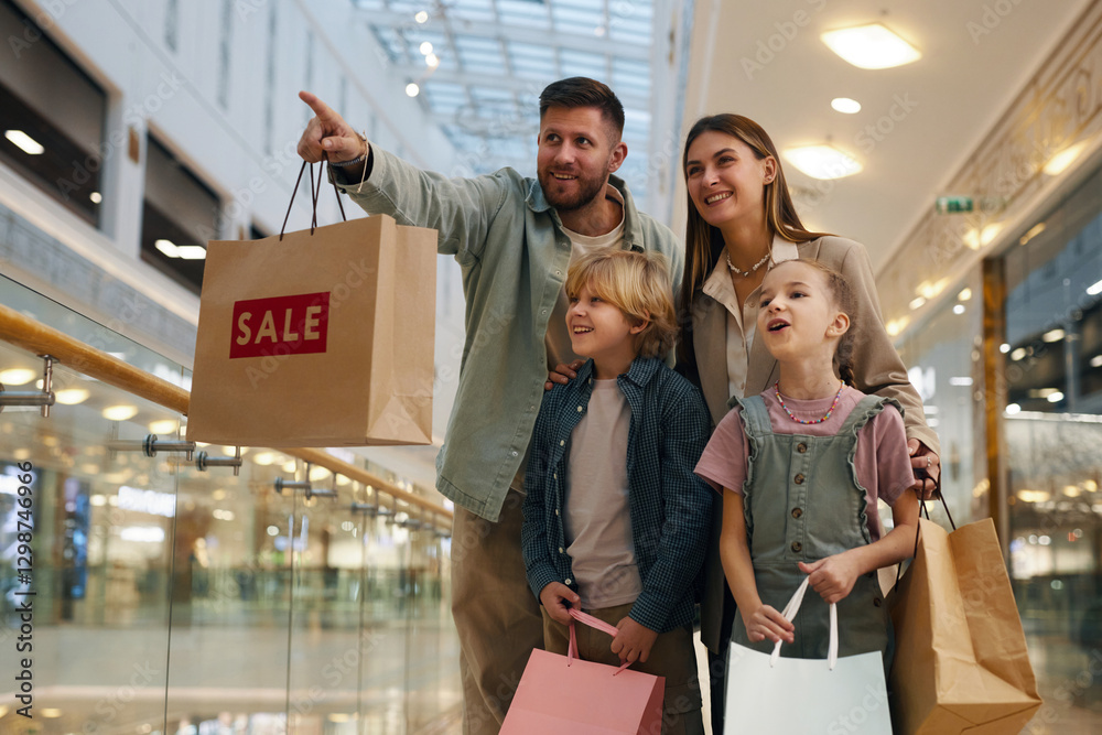 Fototapeta premium Wide shot of young man holding paper bag with sale sign and showing just opened store while his family looking t it with smiles