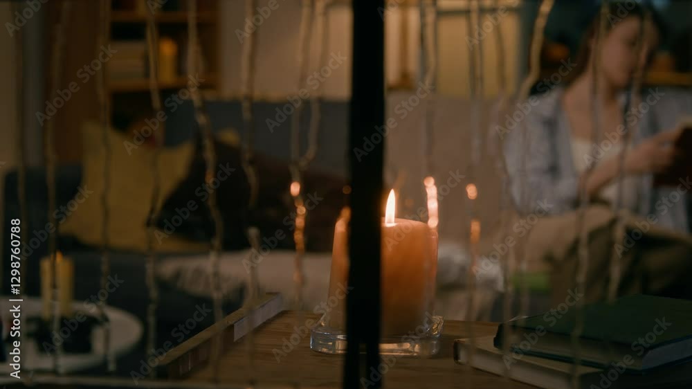 Full panning shot of candle burning on table by window on rainy night, and young woman sitting on couch with blanket, reading book in blurred background, seen by stranger or stalker through window Full panning shot of candle burning on table by window on rainy night, and young woman sitting on couch with blanket, reading book in blurred background, seen by stranger or stalker through window