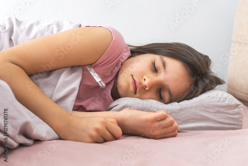 Sick little girl in a pink T-shirt sleeping in bed with a thermometer under her arm. She looks weak and unwell, resting on a pillow. Concept of illness, fever, recovery