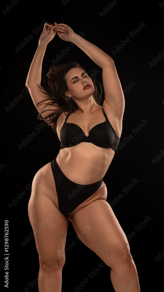 Fototapeta premium Overweight young woman in black underwear, posing in relaxed posture with closed eyes and holding arms raised against black studio background. Concept of natural beauty, self love, confidence.